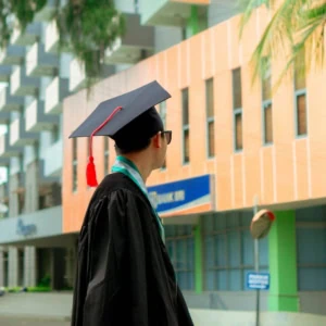 Étudiant diplômé vu de dos en toge noire, toque à pompon rouge, sur campus urbain.