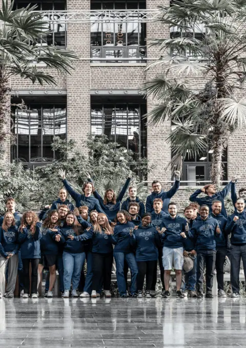 Groupe de jeunes en sweats à capuche devant façade en brique et palmiers.