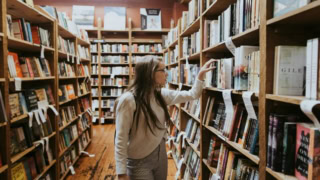 Librairie indépendante, allée de rayonnages en bois, personne feuilletant des livres.