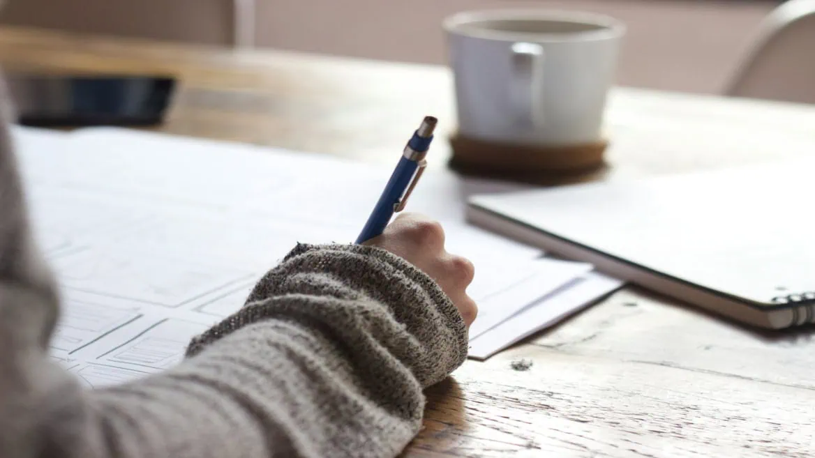 Main tenant un stylo bleu au-dessus desquisse sur table en bois, tasse et carnet flous.