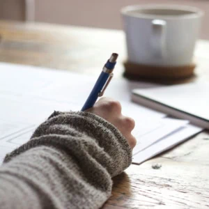 Main tenant un stylo bleu au-dessus desquisse sur table en bois, tasse et carnet flous.