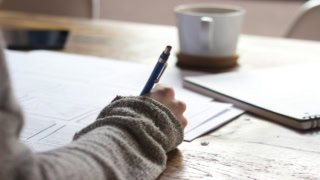 Main tenant un stylo bleu au-dessus desquisse sur table en bois, tasse et carnet flous.