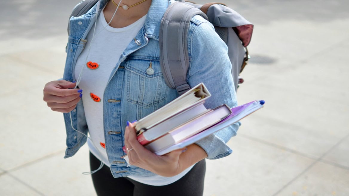 Étudiant(e) marchant avec livres, veste en jean, sac à dos et dossier violet.