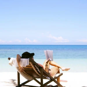 Personne lisant sur chaise longue au soleil sur plage de sable face à la mer.
