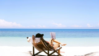 Personne lisant sur chaise longue au soleil sur plage de sable face à la mer.
