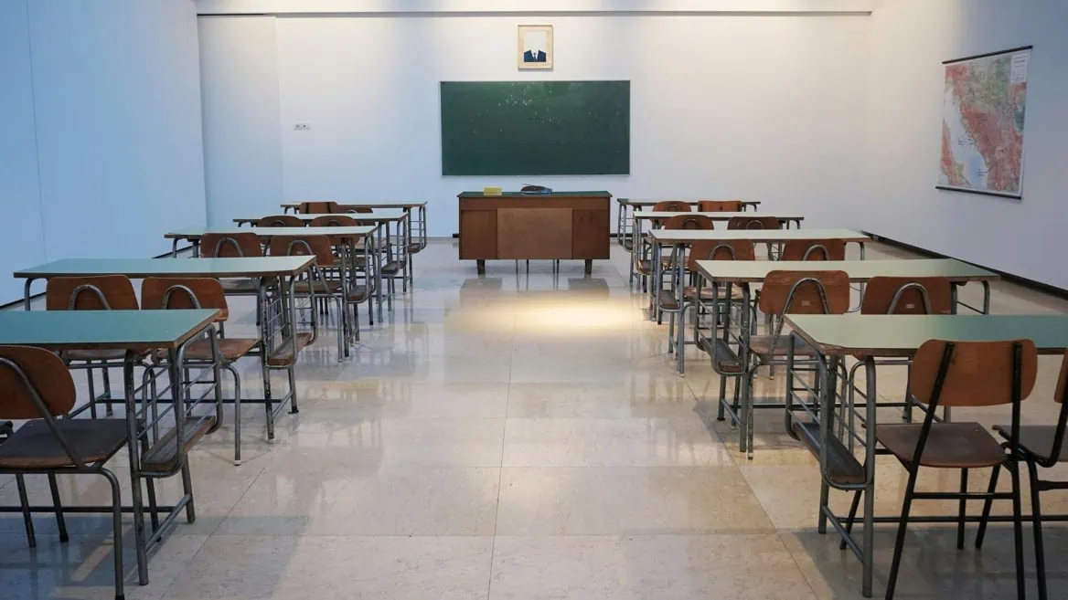 Salle de classe vide symétrique avec rangées de pupitres, bureau du professeur et tableau noir.