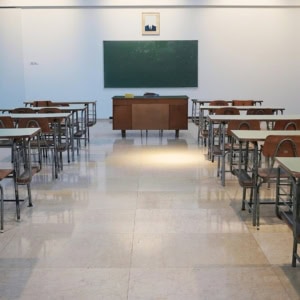 Salle de classe vide symétrique avec rangées de pupitres, bureau du professeur et tableau noir.