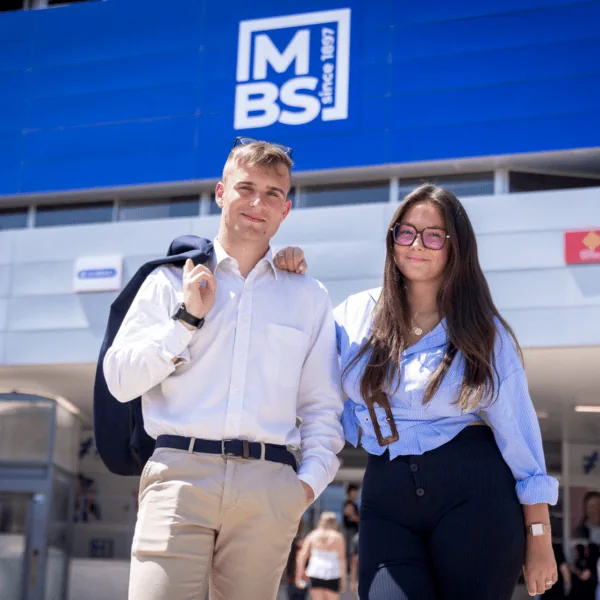 Deux jeunes étudiants posent devant un bâtiment moderne bleu sur un campus lumineux.