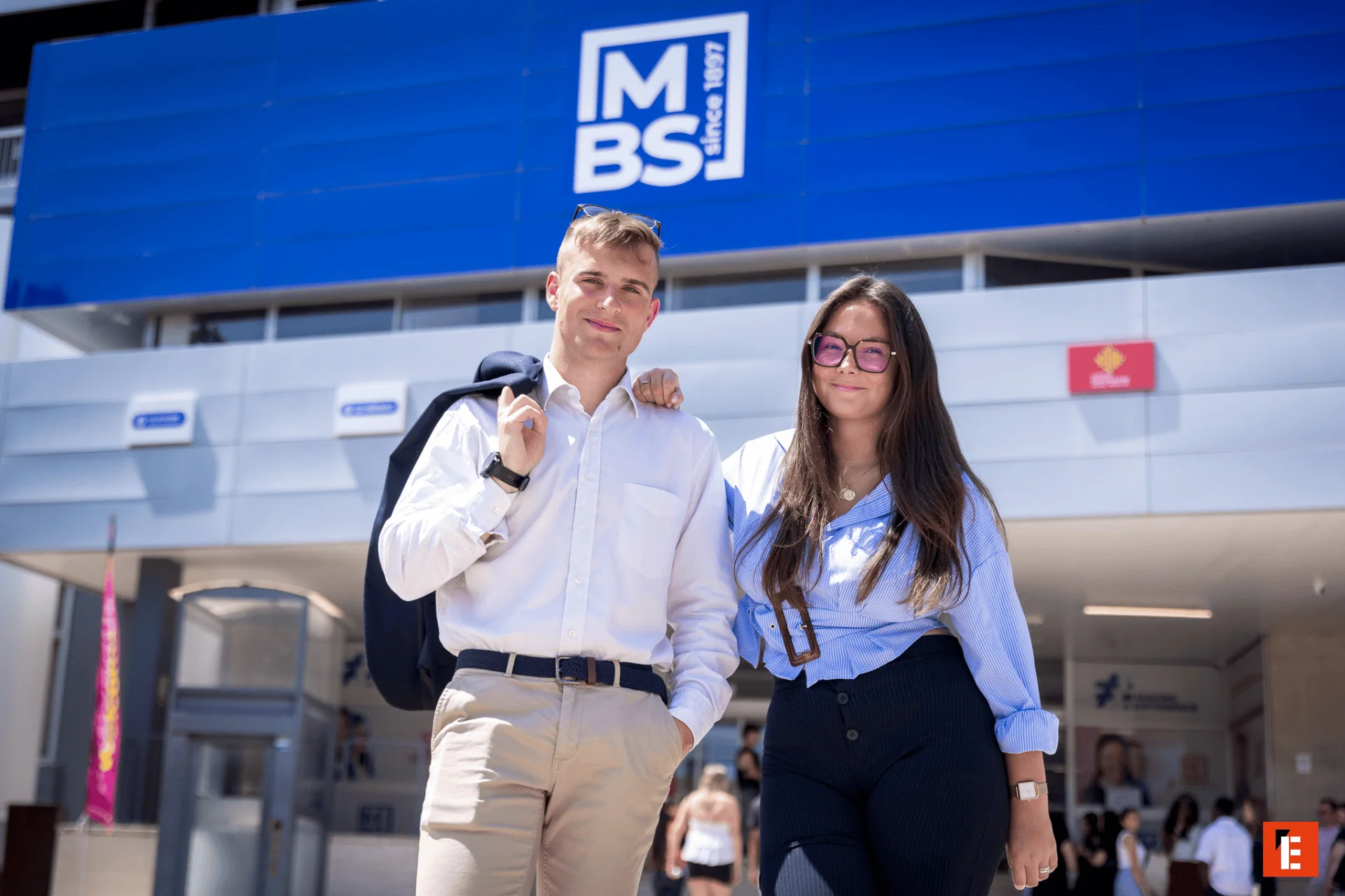 Deux jeunes étudiants posent devant un bâtiment moderne bleu sur un campus lumineux.