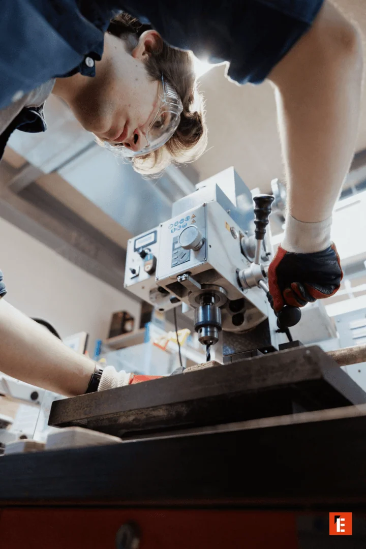 Opérateur en atelier perçage de précision à la perceuse, gants et lunettes de sécurité.
