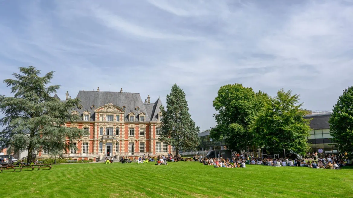 Campus avec pelouse et bâtiment historique en brique, étudiants pique-niquant au soleil.