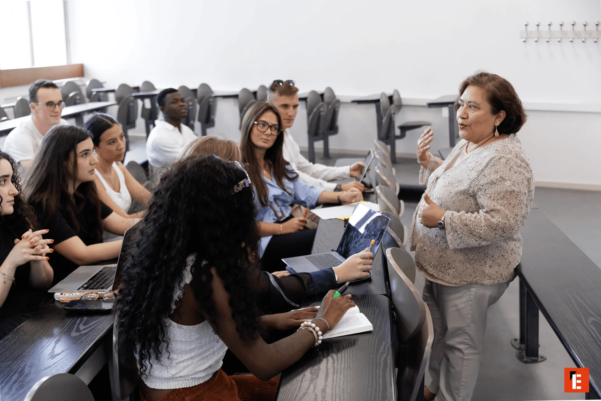 Cours interactif en amphithéâtre, professeure debout et étudiants attentifs prenant des notes.