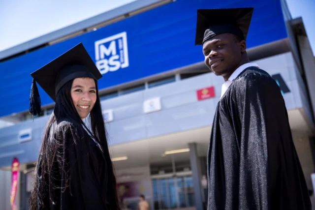 Deux étudiants diplômés en toges et birettes sur campus universitaire moderne.
