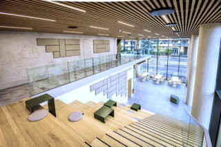 Atrium lumineux avec gradins en bois, plafond à lamelles, verrière et mobilier minimaliste.