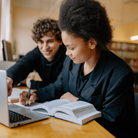 Étudiants en révision avec livre et ordinateur dans une bibliothèque chaleureuse, travail collaboratif.