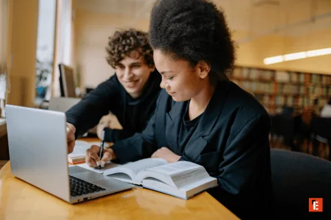 Étudiants en révision avec livre et ordinateur dans une bibliothèque chaleureuse, travail collaboratif.