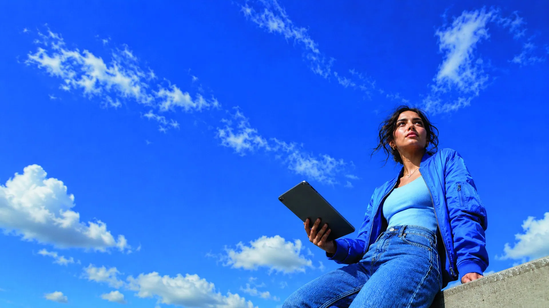 Femme assise en veste bleue tenant une tablette sous un vaste ciel bleu.