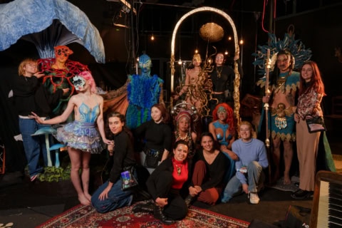 Groupe en coulisses avec costumes théâtraux, maquillage, sculptures et artisans sur scène.