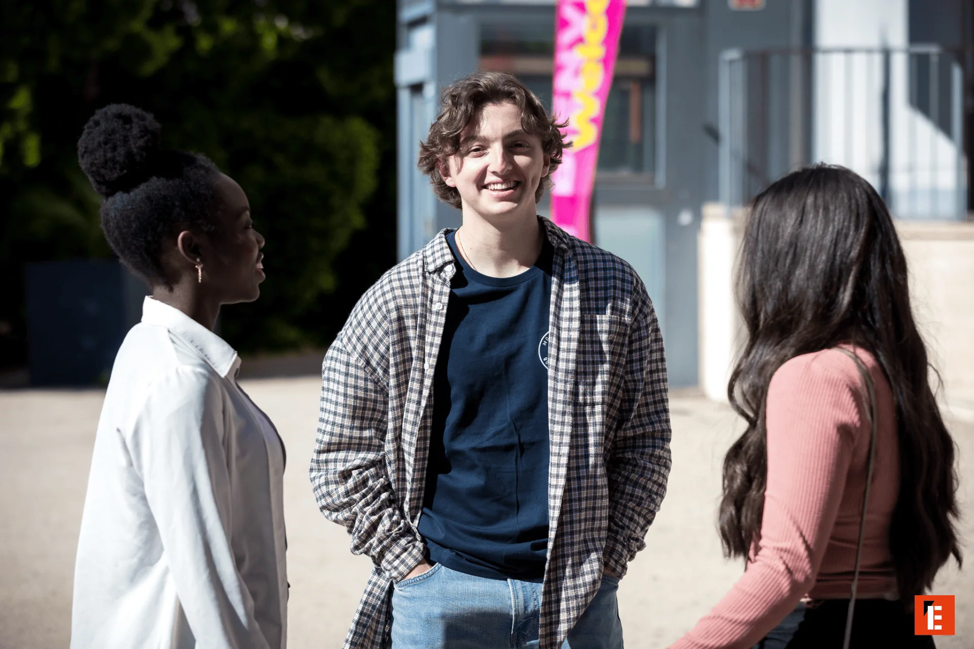 Trois jeunes amis discutent en extérieur au soleil, ambiance décontractée et urbaine.