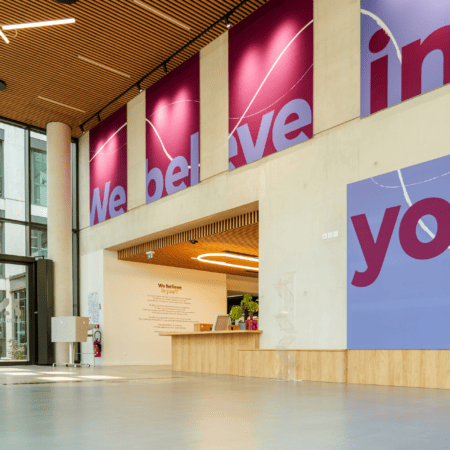 Hall daccueil moderne lumineux avec réception, bois, béton et bannières magenta et pervenche.