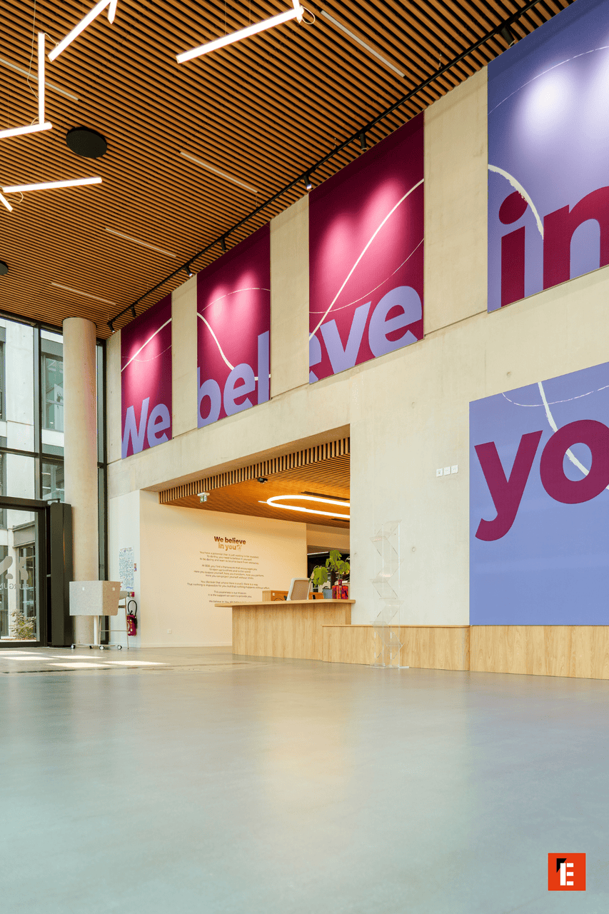 Hall daccueil moderne lumineux avec réception, bois, béton et bannières magenta et pervenche.