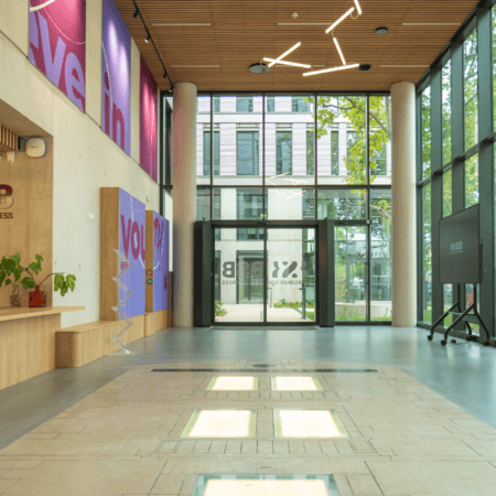 Lobby moderne lumineux avec façade vitrée, double hauteur, plafond bois et sol carrelé éclairé.