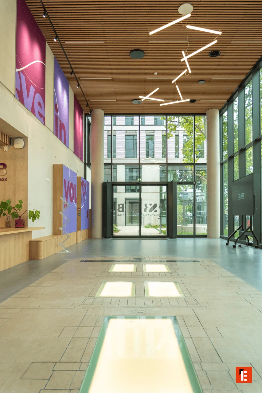 Lobby moderne lumineux avec façade vitrée, double hauteur, plafond bois et sol carrelé éclairé.