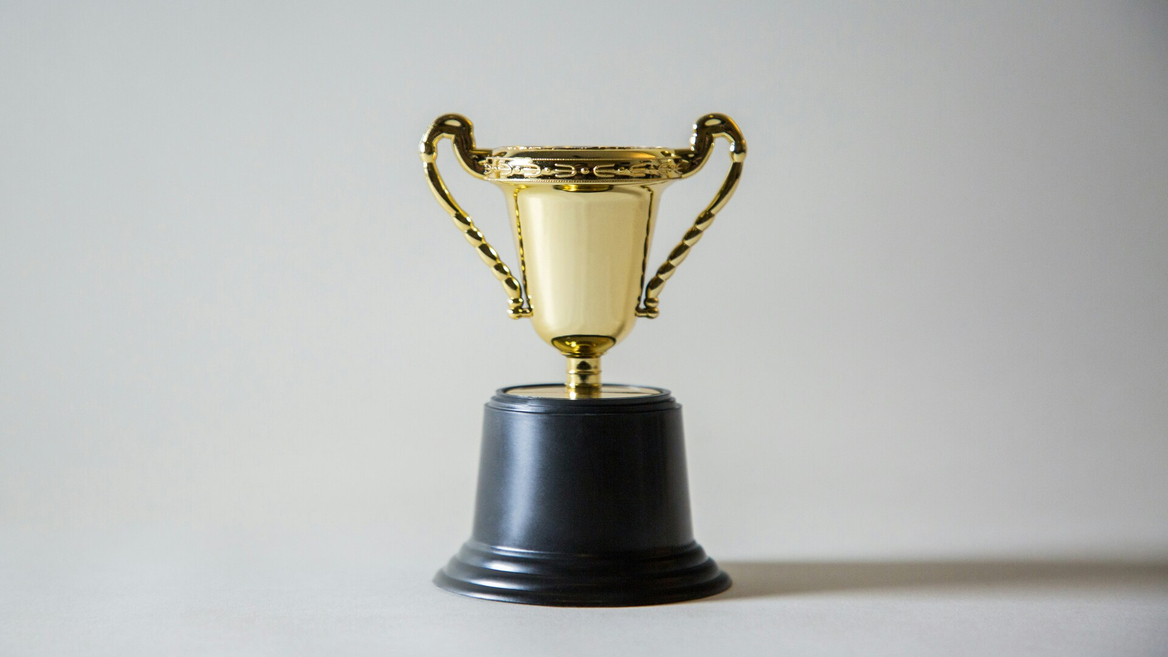 Petit trophée en or, coupe sur socle noir, fond neutre.
