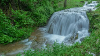 Cascade forestière, rivière en longue exposition, eau lisse, végétation verte, pierres couvertes de mousse.