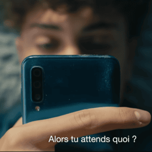 Smartphone bleu tenu à lhorizontale par une main, visage flou, texte «Alors tu attends quoi ?».