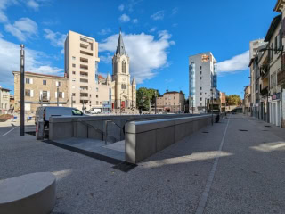 Place urbaine piétonne avec église à flèche, bâtiments modernes, commerces et ciel bleu.