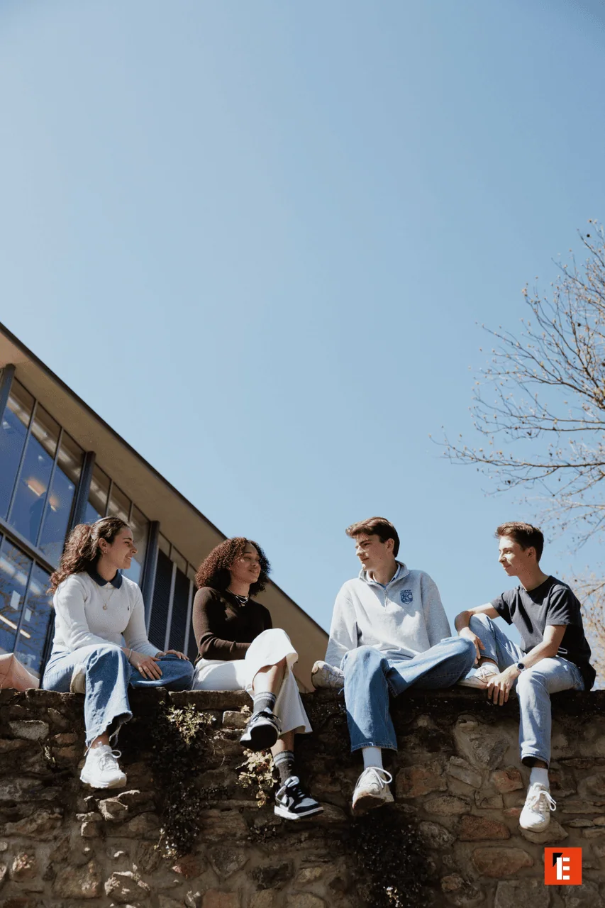 Quatre étudiants assis sur un mur de campus, discutant sous un ciel bleu printanier.