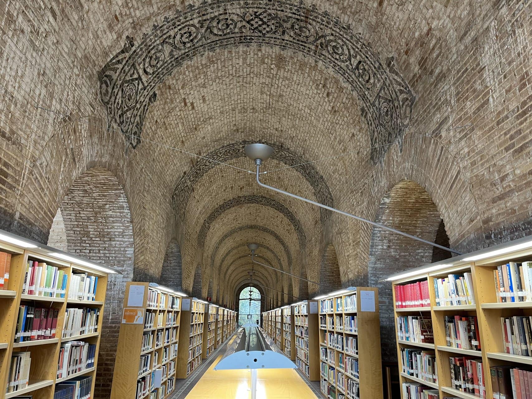 Salle de lecture voûtée en brique, longues étagères en perspective et lumière naturelle.