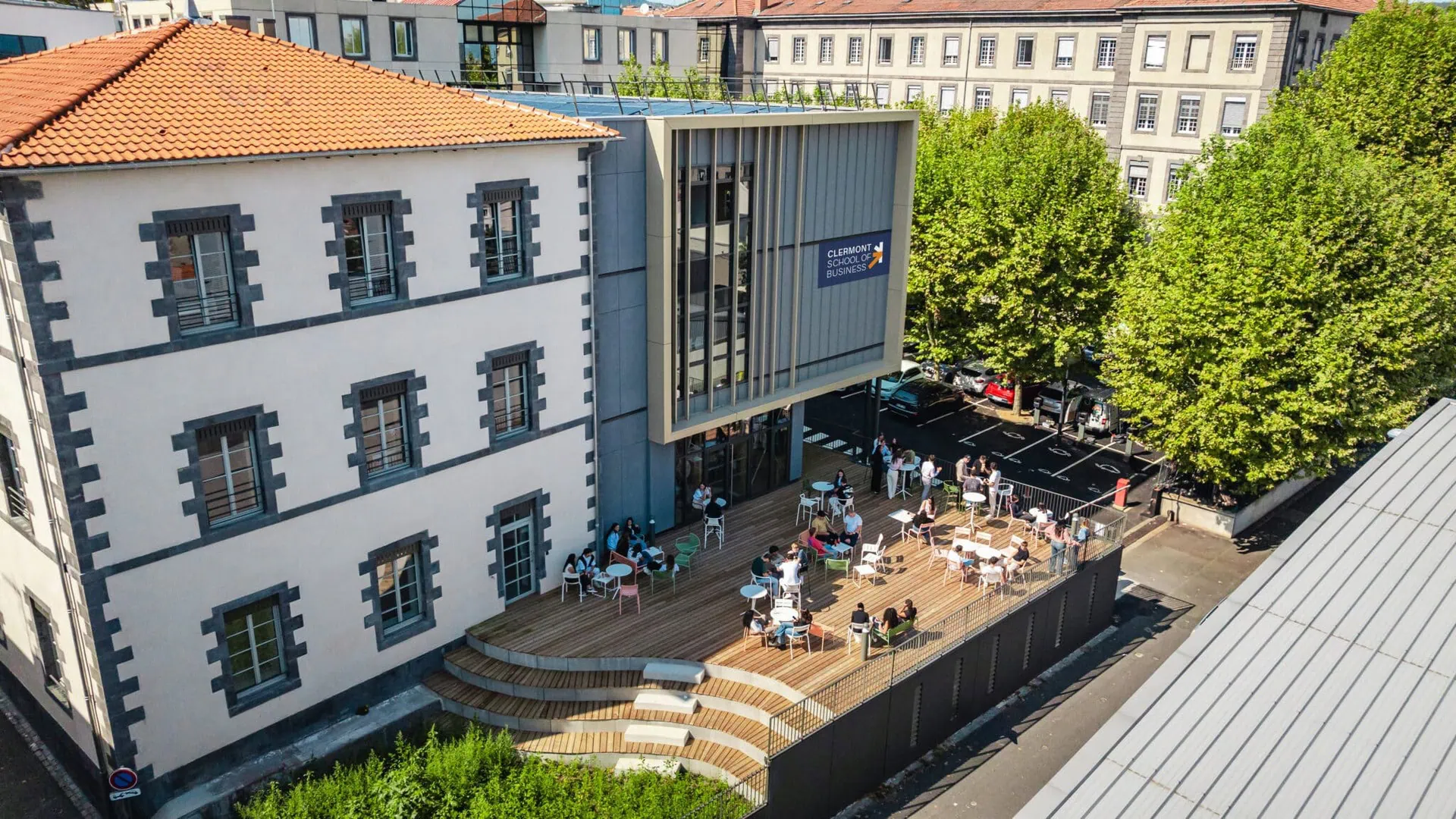 Terrasse bois animée du campus Clermont École de commerce, bâtiment ancien et annexe moderne.