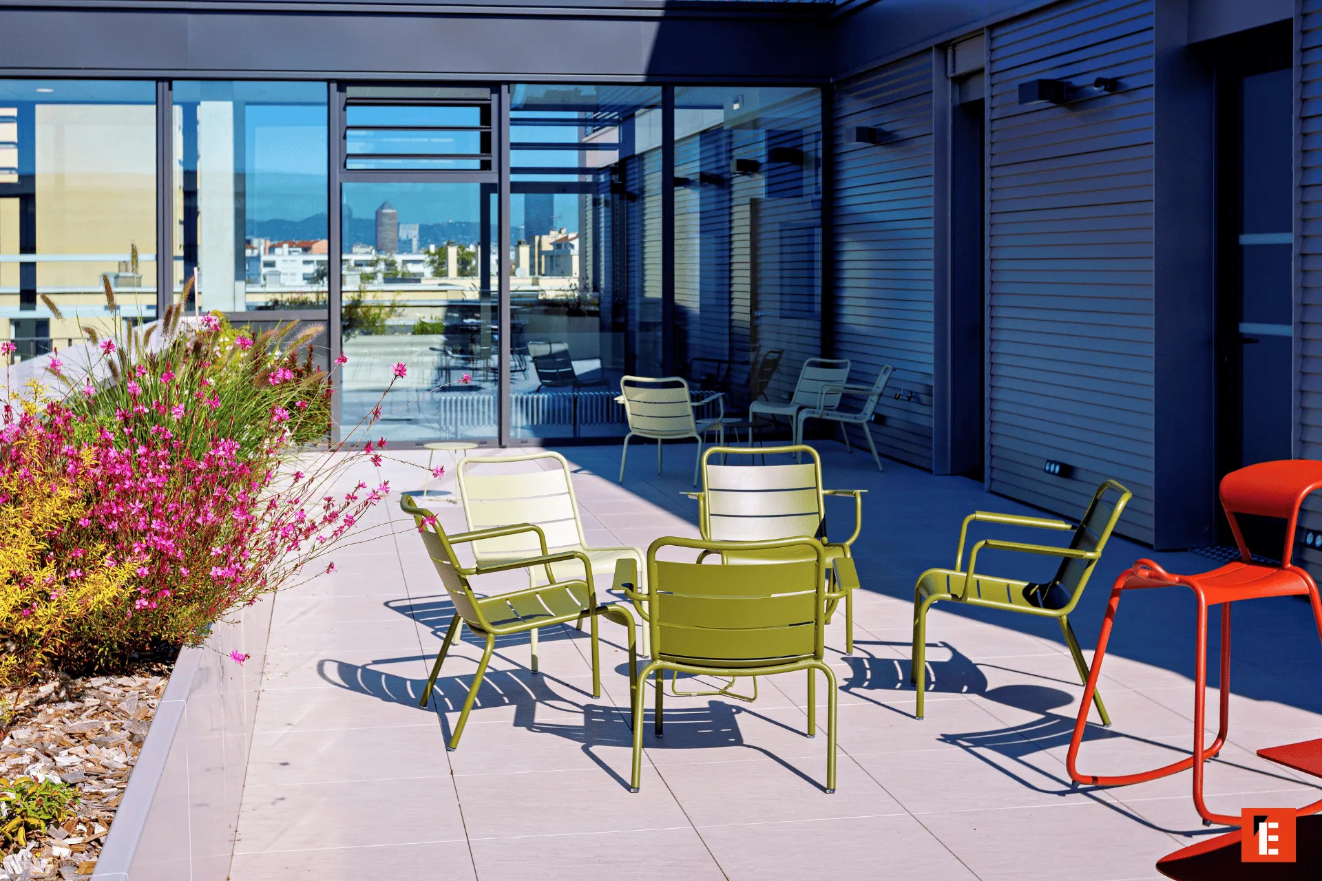 Terrasse sur toit moderne avec mobilier vert olive, tabourets rouges, plantes et vue urbaine.