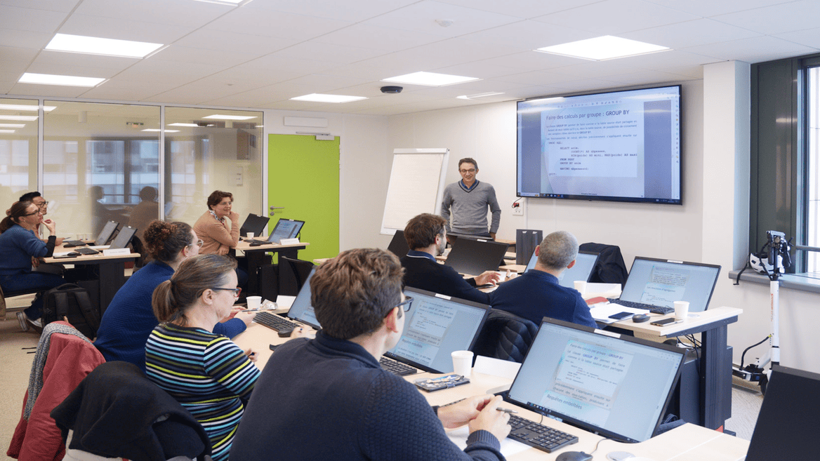 Salle de formation informatique avec présentateur, participants et ordinateurs affichant cours SQL.