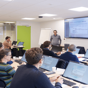 Salle de formation informatique avec présentateur, participants et ordinateurs affichant cours SQL.