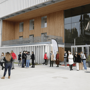 Bâtiment contemporain en verre, bois et métal, foule devant lentrée.