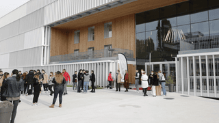 Bâtiment contemporain en verre, bois et métal, foule devant lentrée.