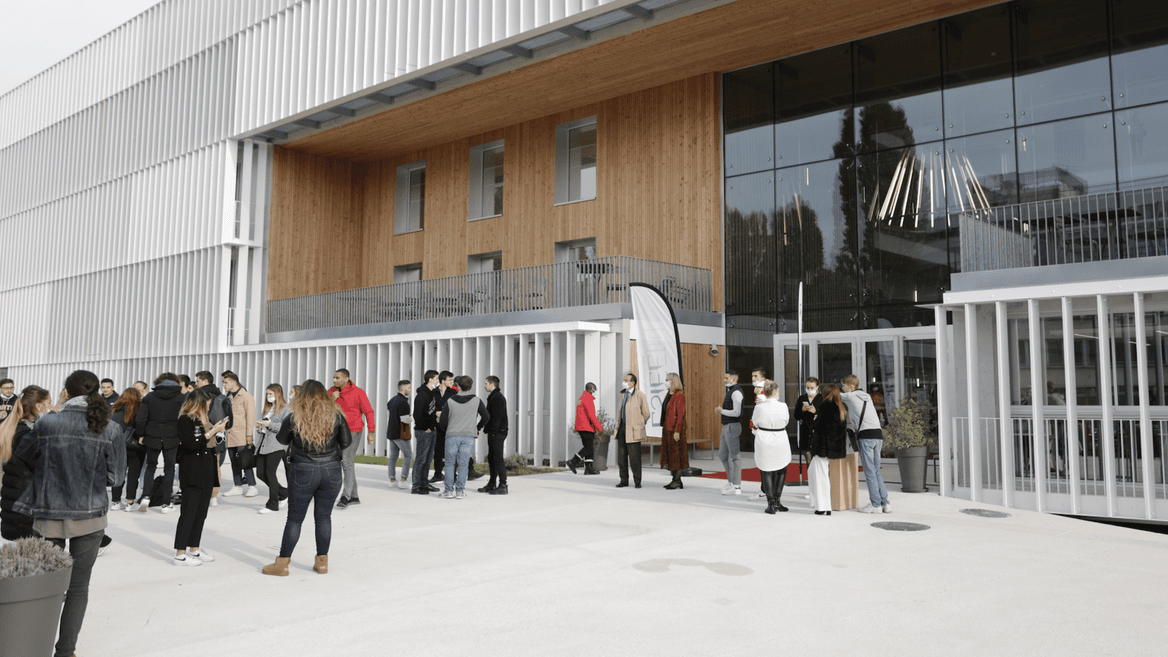 Bâtiment contemporain en verre, bois et métal, foule devant lentrée.