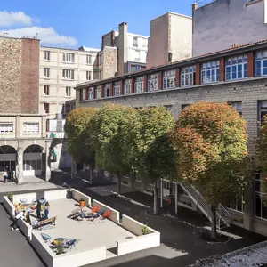 Cour urbaine ensoleillée entourée de façades modernes, arbres, terrasse et chaises colorées.