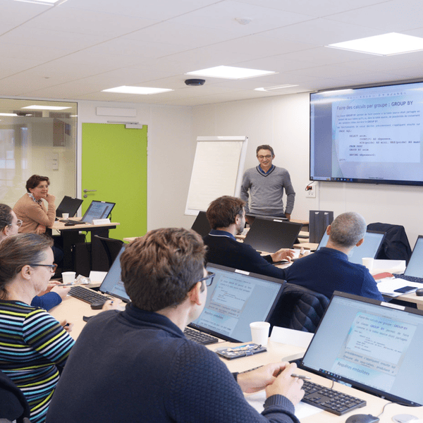 Salle de formation informatique avec présentateur, participants et ordinateurs affichant cours SQL.