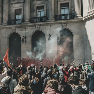 Manifestation devant monument classique à colonnes, foule dense, fumée colorée et drapeaux rouges.