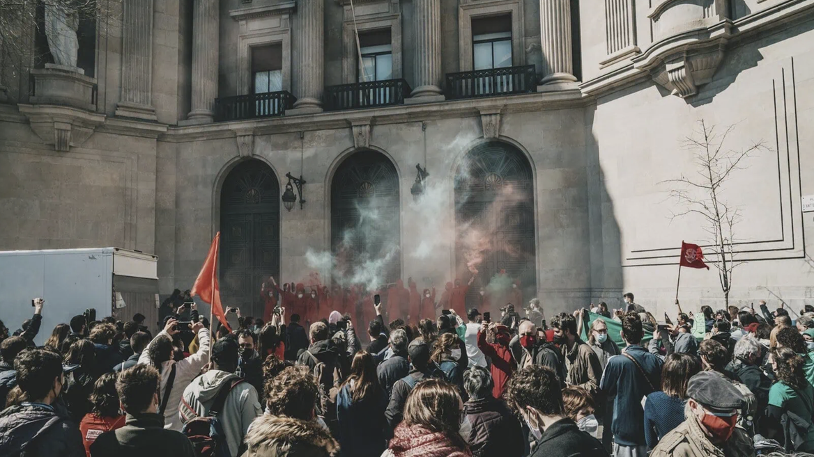 Manifestation devant monument classique à colonnes, foule dense, fumée colorée et drapeaux rouges.