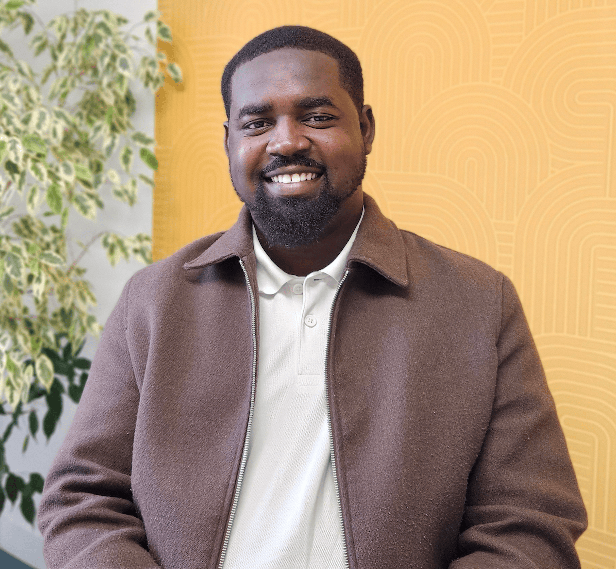 Portrait professionnel dhomme souriant en polo et veste marron, fond jaune graphique avec plante.