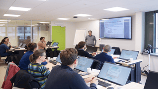 Salle de formation informatique avec présentateur, participants et ordinateurs affichant cours SQL.